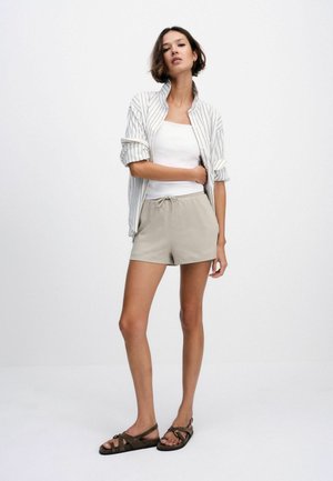 Frau steht mit der Hand in der Tasche, trägt beige Shorts mit Kordelzug, ein weißes Tanktop, ein gestreiftes Hemd mit hochgekrempelten Ärmeln und braune Sandalen.