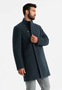 Cappotto in lana blu navy con colletto alto, dotato di due bottoni, taglio dritto e tasche laterali. Texture liscia e design su misura.