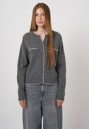 Jonge vrouw met lang steil haar die een grijze gebreide cardigan met witte rand en losse grijze spijkerbroek draagt, staand tegen een effen achtergrond.
