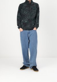 Osoba nosząca czarny i zielony wzorzysty polar, jasnoniebieskie luźne dżinsy i czarne trampki z gumowymi podeszwami, ręce w kieszeniach.