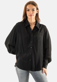Camisa negra de manga larga con cuello clásico, con botones en el frente y detalles de encaje en el escote. Tejido suave con un corte holgado y un bolsillo en el pecho.