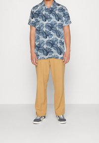 Camicia tropicale azzurra a maniche corte con stampa a foglie, abbinata a pantaloni chiari color sabbia e scarpe blu navy con dettagli bianchi.