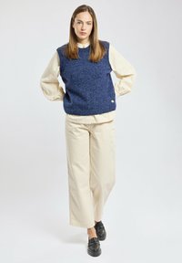 Granatowa wełniana kamizelka o teksturowanym wykończeniu, noszona na kremowej koszuli. W połączeniu z kremowymi spodniami o szerokich nogawkach oraz czarnymi mokasynami.