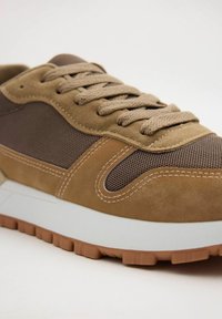 Zapatillas de ante marrón y malla con un upper texturizado, cordones redondos y una mediasuela blanca. Cuenta con una suela de goma con un dibujo en la banda de rodadura.