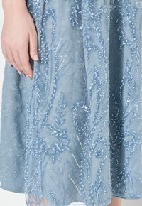 Gebreide blauwe jurk met ingewikkelde bloemenborduursels, gelaagde texturen en een ontwerp tot op de knie. Hand rust op de stof.