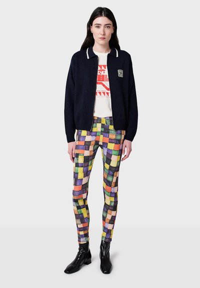 Marinblå stickad bomberjacka med vita krage detaljer, vit t-shirt med grafik under, och flerfärgad mönstrad legging. Svarta ankelskor.