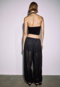 Crni top bez naramenica u kombinaciji s prozirnim crnim hlačama širokih nogavica s uzdužnim prugama i teksturiranim završetkom, gledano odostraga.