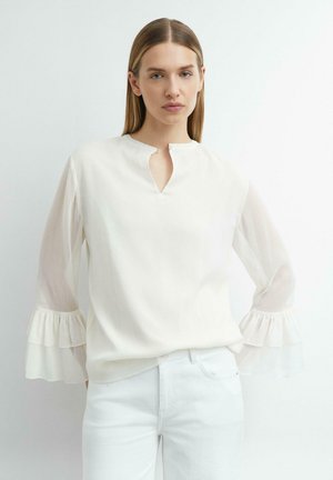 Vrouw draagt een wit blouse met geplooide doorschijnende mouwen en een kleine voorscheur aan de halslijn, gecombineerd met een witte broek, staand tegen een effen achtergrond.
