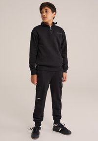 Melnā džemperis ar ceturtdaļas rāvējslēdzēju un ribotām piedurknēm, savienots ar melniem kravas biksēm ar sānu kabatām un logotipu. Strukturēta auduma.