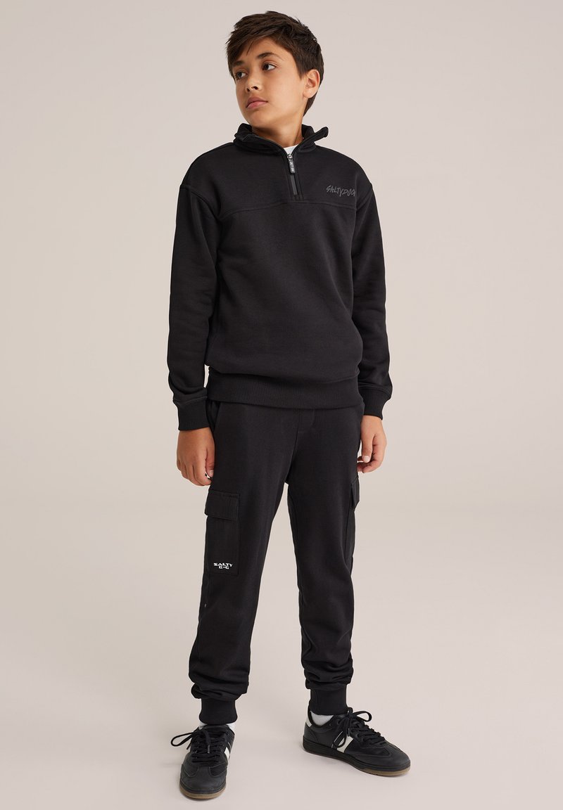 Hanorac negru cu fermoar la jumătate și manșete cu talie, purtat cu pantaloni cargo negri, dotati cu buzunare laterale și un logo. Material texturat.