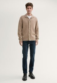 Beige Strickjacke mit Schalkragen, sieben Knöpfen und Rippbündchen, kombiniert mit dunkelblauen Jeans und schwarzen Lederstiefeln.