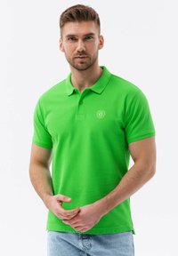 Zelené polo tričko s žebrovanou texturou, krátkými rukávy a zapínáním na tři knoflíky, s malým logem na levé části hrudi.