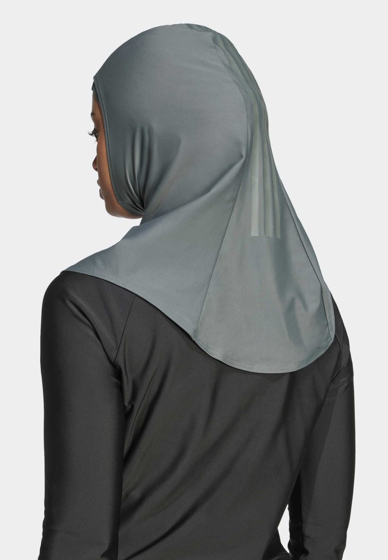 Frau, die ein hellgraues Sport-Hijab mit dezenten vertikalen Streifen trägt, kombiniert mit einem schwarzen Langarmtop und nach links blickt.