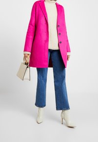 Cappotto in feltro fucsia con una vestibilità sartoriale, revers a banda, e tre bottoni, abbinato a un maglione crema, jeans a zampa d'elefante blu e tacchi alti bianchi.