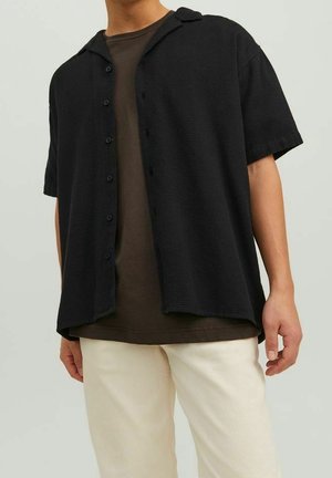 Černá krátká košile s knoflíky z texturované látky s límečkem ve stylu kempu. Nositelná přes tmavě hnědou košili, k tomu béžové kalhoty.