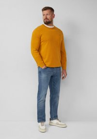 Mosterdgele trui met ronde hals en lange mouwen, gecombineerd met lichtblauwe spijkerbroek en witte sneakers met tan accenten.
