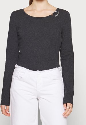 Femme portant un haut ajusté à manches longues gris foncé avec des boutons et une broderie près de l'encolure, associé à un pantalon blanc taille haute.