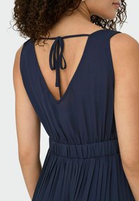 Navy blue dress featuring a pleated design, deep V-back, and adjustable tie closure. Soft fabric with a smooth texture.