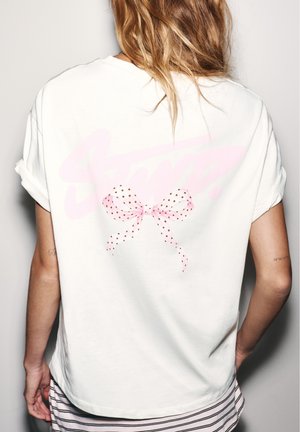 Personne aux cheveux châtain clair porte un t-shirt blanc avec manches retroussées et un nœud rose à pois sur le dos, associé à un bas rayé.