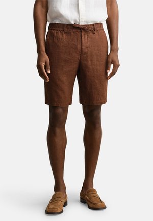 Mand iført brune linnedshorts med forlommer og snøre i taljen, kombineret med brune loafers og en hvid skjorte.