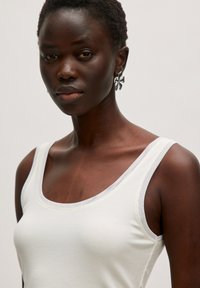 Weißes, geripptes Tanktop mit einem runden Ausschnitt. Das Model trägt einen dekorativen schwarzen Ohrring. Der Hintergrund ist neutral und schlicht.