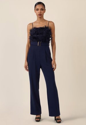 Frau trägt navyblaues ärmelloses Feder-Top und weite, taillierte Hosen mit Gürtel sowie schwarze Plateau-Absatzschuhe, steht vor schlichtem beige Hintergrund.