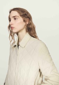 Světlá béžová prošívaná bunda s diamantovým vzorem, která má límec a zapínání na knoflíky. Materiál vypadá hladce a má měkkou texturu.