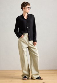 Personne avec les cheveux courts et des lunettes de soleil portant une chemise noire à boutons, un pantalon beige large et des sandales noires, debout sur un sol en bois.