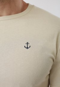 Κοντινό πλάνο ενός ατόμου που φοράει μπεζ μπλουζάκι με ένα μικρό κεντημένο ναυτικό μπλε άγκυρα στο στήθος.