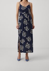 Kvinna i en marinblå maxiklänning med vita blommönster, tunna axelremmar, sidoklyvning och matchande bälte, tillsammans med blå spetsiga klackskor.