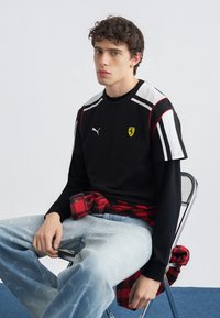 Mlad moški s kodrastimi lasmi sedi na kovinskem stolu, oblečen v črno majico Puma Ferrari, podloženo z dolgo majico, in svetlo modre kavbojke, rdečo karo srajco pa ima zavezano okoli pasu.