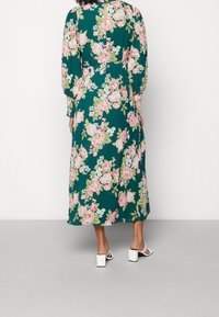 El vestido floral presenta un fondo verde con grandes rosas rosas y acentos blancos. Tiene mangas largas y una cintura ajustada, con un flujo que llega hasta la mitad de la pantorrilla.