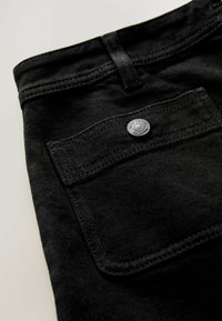 Schwarzer Denim-Stoff im Detail, der eine Nahaufnahme einer Tasche, eines Metallknopfs und der Naht zeigt. Glatte Textur mit feinem Fischgrätenmuster.