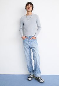 Lys grå ribbet langermet skjorte, blå mønstrede jeans og grå joggesko med svarte accenter. Modellen står mot en hvit vegg.