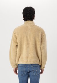 Chaqueta de forro polar beige con cuello alto, corte holgado, textura suave y puños acanalados, combinada con unos vaqueros de mezclilla azul. Sin logotipos visibles.