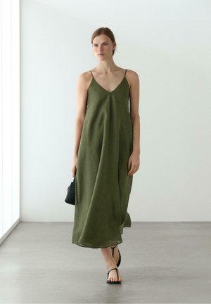 Donna che cammina al chiuso indossando un abito lungo verde oliva senza maniche, sandali neri e tenendo una piccola borsa nera.