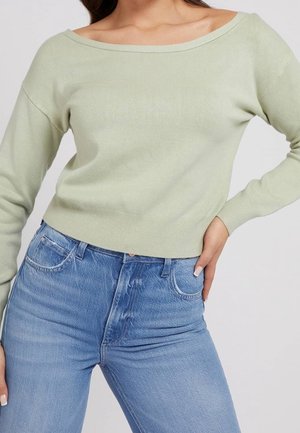 Femme portant un pull vert clair épaules dénudées et un jean bleu taille haute, main posée sur la hanche sur un fond uni.
