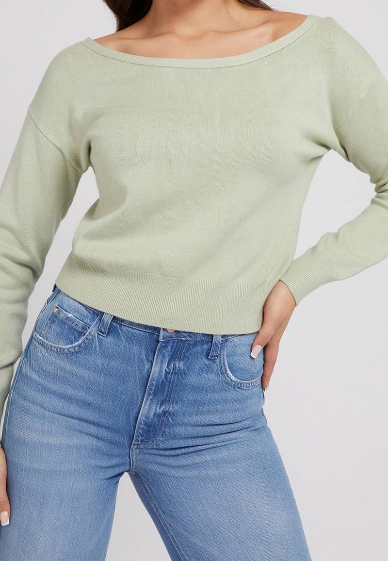 Femme portant un pull vert clair épaules dénudées et un jean bleu taille haute, main posée sur la hanche sur un fond uni.