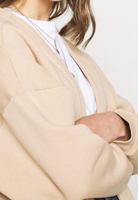 Cárdigan beige de gran tamaño con mangas tipo globo, textura suave, apertura delantera, superpuesto sobre una camiseta blanca, mostrando un ajuste relajado.