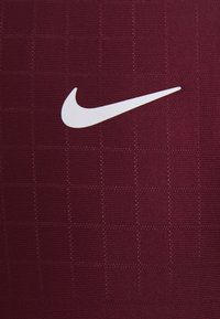 Camiseta deportiva color marrón oscuro hecha de tela texturizada, con un logo de swoosh blanco de Nike y un patrón de cuadrícula en el material.