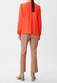 Blusa lunga a maniche corte di un vivace arancione con maniche a sbuffo, abbinata a pantaloni a quadri marroni e stivaletti beige con tacco.