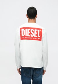 Camisa de manga longa branca com o logótipo vermelho "DIESEL" e o texto "PARA UMA VIDA DE SUCESSO" nas costas. Corte casual, material de algodão, design de gola redonda.