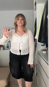 Bjeli pleteni kardigan s valovitim rubom i gumbima, nosi se preko bijelog topa i uparuje s crnim širokim hlačama s detaljima na zatvaraču.