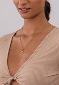 Goldkette mit einem kleinen Herzanhänger und einem blauen Akzentstein, getragen über einem beigefarbenen gerippten Oberteil, das ein elegantes Design zeigt.
