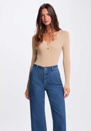 Donna che indossa una maglia a coste beige a maniche lunghe con bottoni e jeans blu a vita alta e taglio ampio, in piedi su sfondo bianco neutro.
