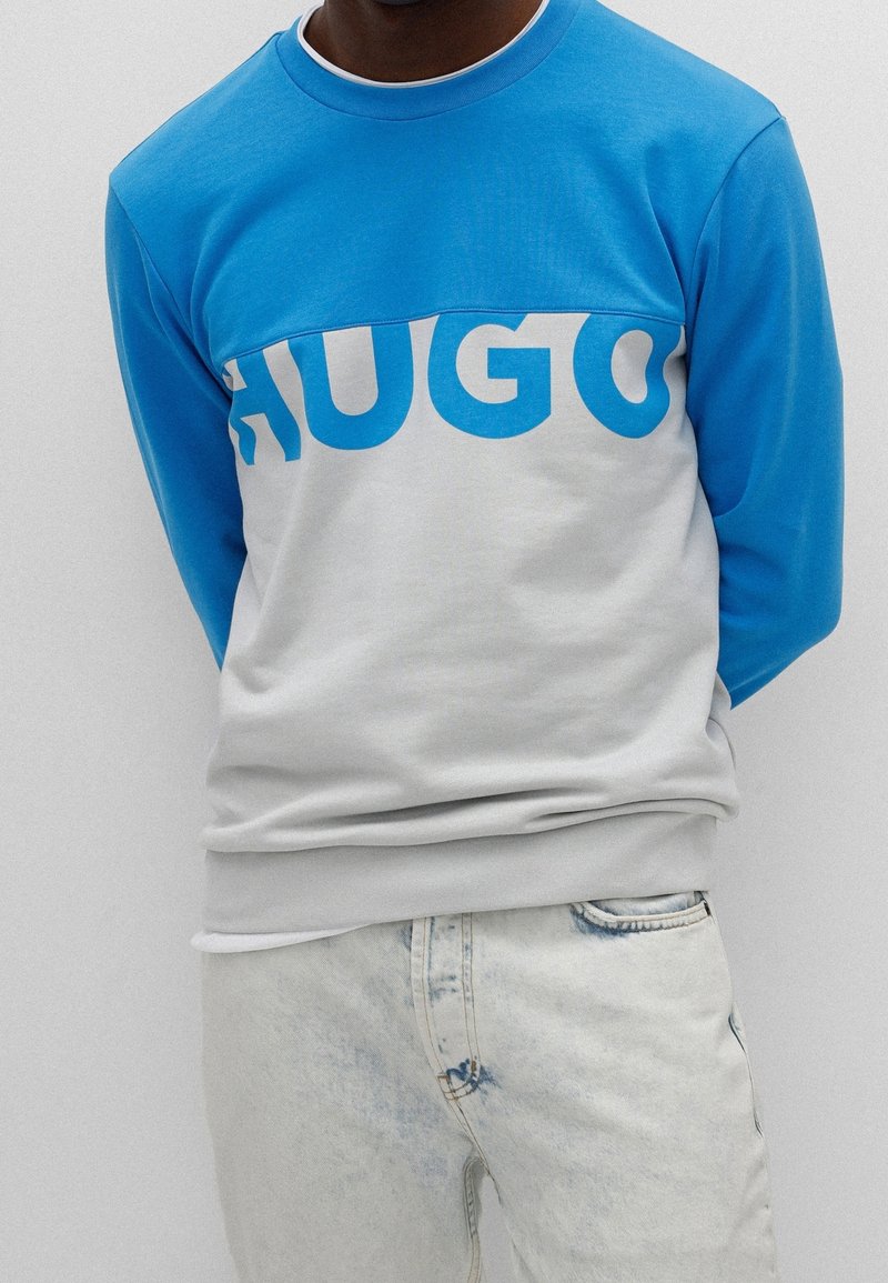 Blau und grauer Kapuzenpullover mit Rundhalsausschnitt, der mit fettem blauen "HUGO"-Text auf der Brust versehen ist, kombiniert mit hellblauen Ärmeln.