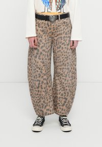 Άτομο που φορά χαλαρά παντελόνια με λεοπάρ μοτίβο, μαύρη ζώνη με αγκράφα σε σχήμα καρδιάς, λευκό μακρυμάνικο μπλουζάκι με γραφικά και μαύρα αθλητικά παπούτσια με λευκά κορδόνια.