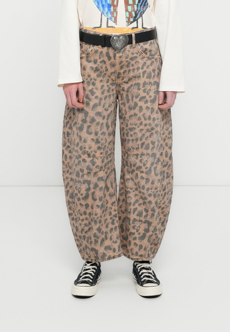 Άτομο που φορά χαλαρά παντελόνια με λεοπάρ μοτίβο, μαύρη ζώνη με αγκράφα σε σχήμα καρδιάς, λευκό μακρυμάνικο μπλουζάκι με γραφικά και μαύρα αθλητικά παπούτσια με λευκά κορδόνια.