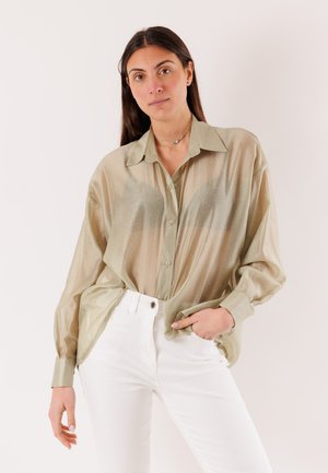 Donna con capelli lunghi e scuri che indossa una camicia beige trasparente abbottonata e pantaloni bianchi, in piedi con una mano nella tasca.