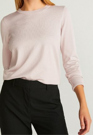 Femme portant un haut rose clair à manches longues, rentré dans un pantalon noir taille haute, sur un fond uni.